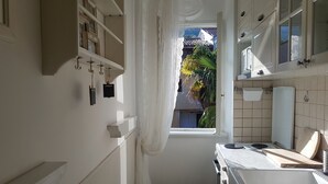A narrow kitchen with white cabinets, a stove, and a window leading to a balcony with a view of palm trees. - Apartment Vesna (Lovran)