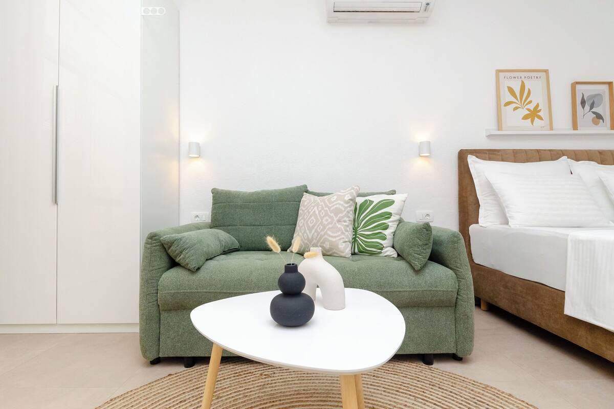 A modern living room with a green sofa, a white coffee table, and a bed with white bedding. There are two wall-mounted lights and framed artwork above the sofa.