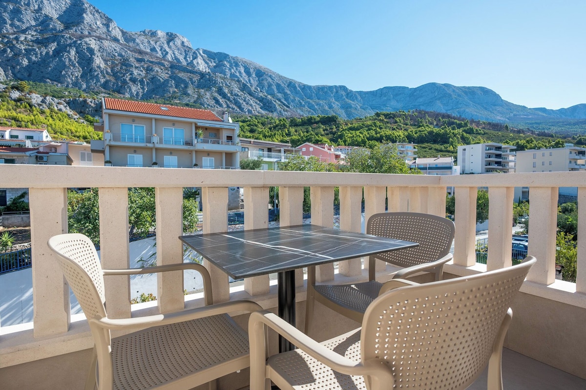 A balcony with a table and chairs, overlooking a mountainous landscape with buildings in the background.
