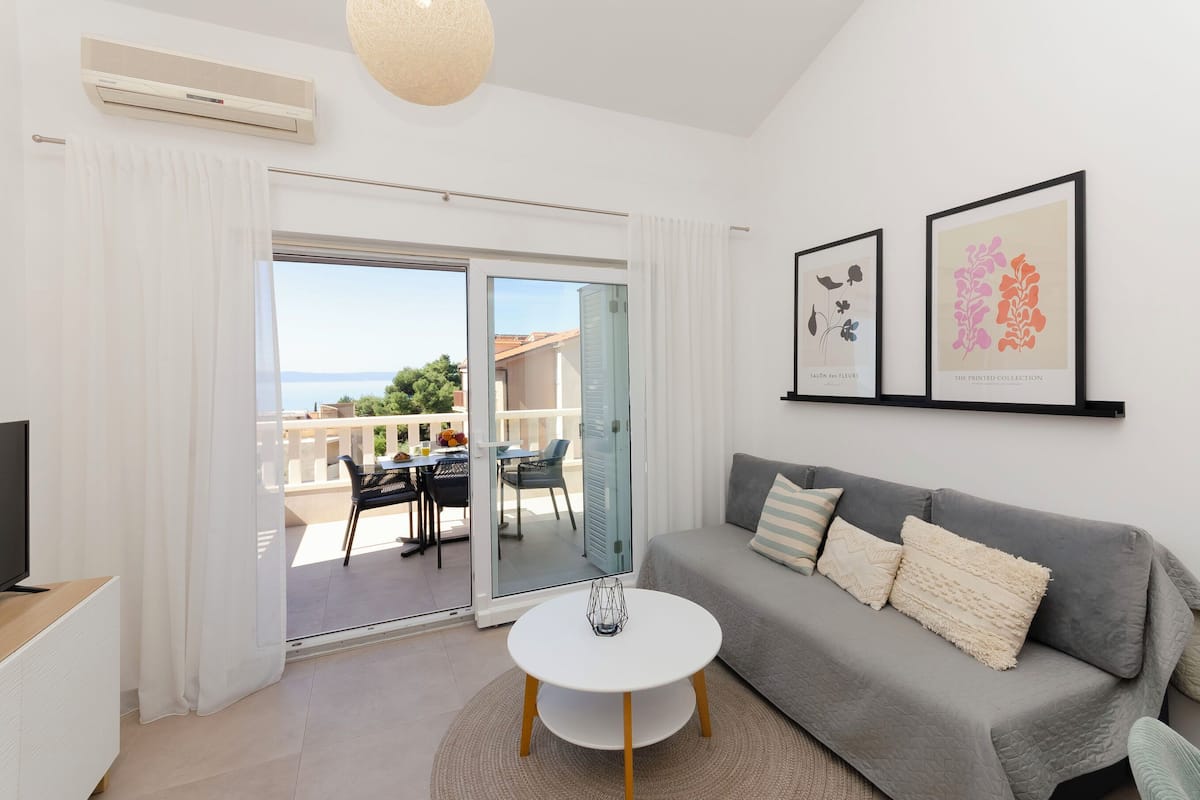 A modern living room with a grey sofa, a white coffee table, and a TV on a wooden stand. There are two framed artworks on the wall and a balcony with a view of the sea.