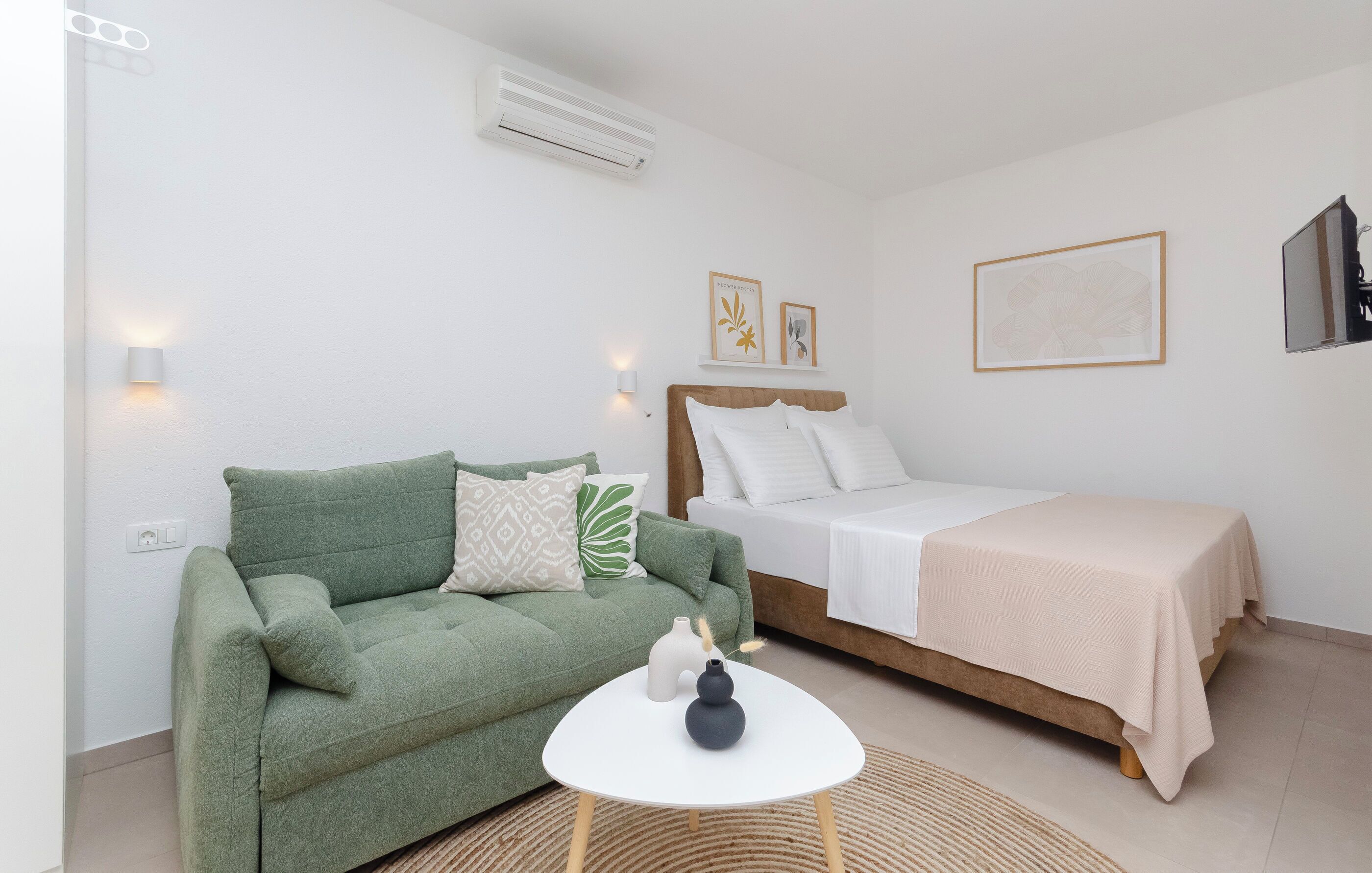A room with a green sofa, a bed with a white bedspread, a small round table with two vases, and wall-mounted artwork.