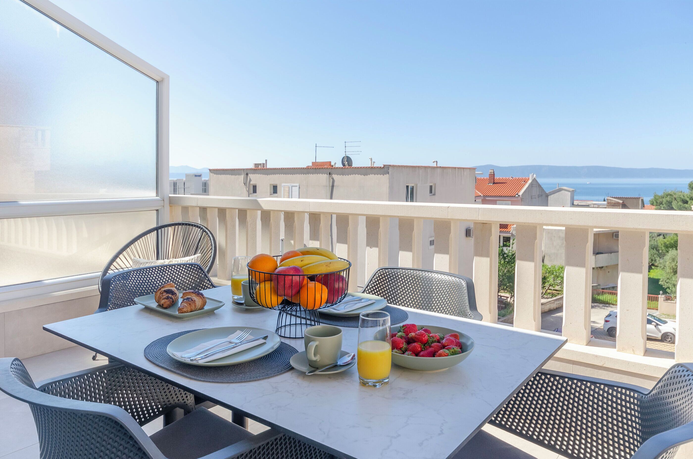 A balcony with a table set for breakfast, including croissants, fruit, and juice, overlooking a cityscape.