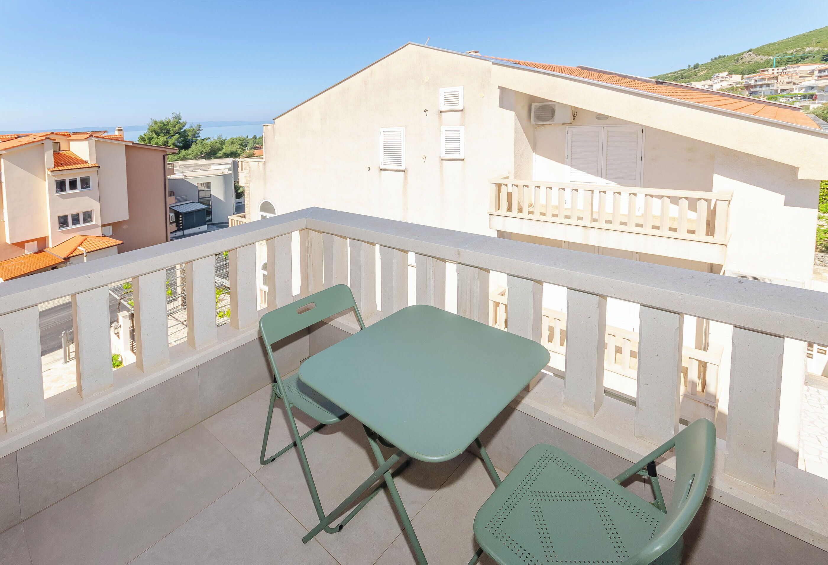 A balcony with a table and two chairs, overlooking a residential area with buildings and a clear sky.