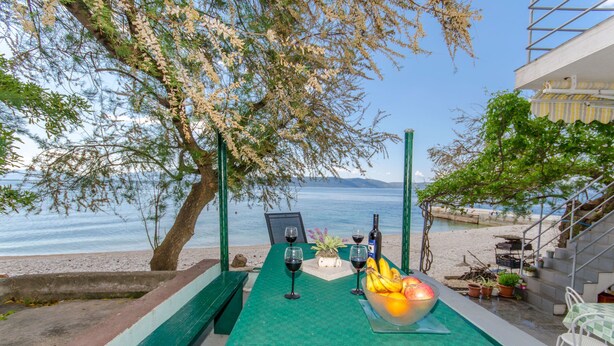 An outdoor seating area with a table set for two, a bowl of fruit, and a bottle of wine. - House Beach House Toma (Zivogosce)