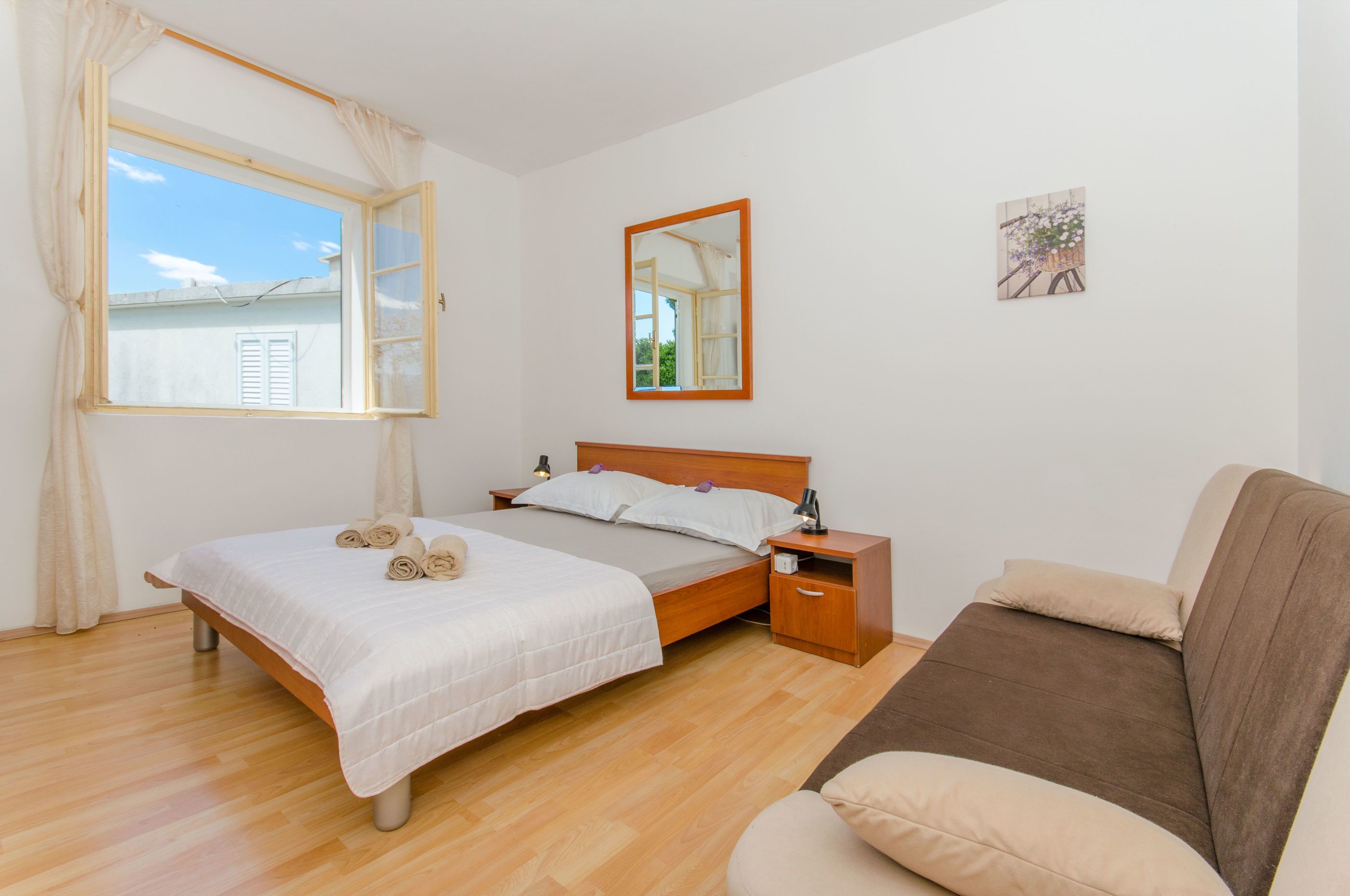 A bedroom with a wooden bed, a nightstand, a bedside table, a window with curtains, and a wall-mounted mirror.