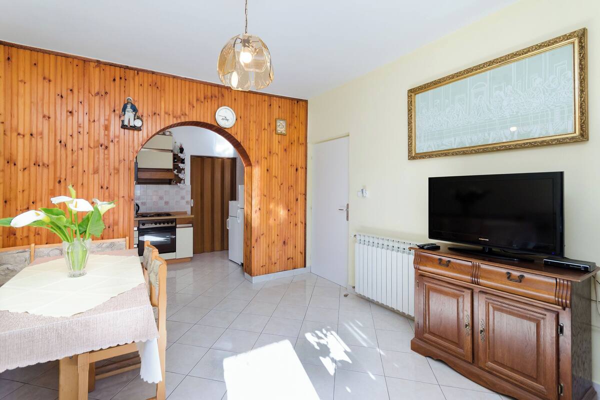 A kitchen with wooden walls, a dining table with a vase of flowers, a TV on a wooden cabinet, and a clock on the wall.