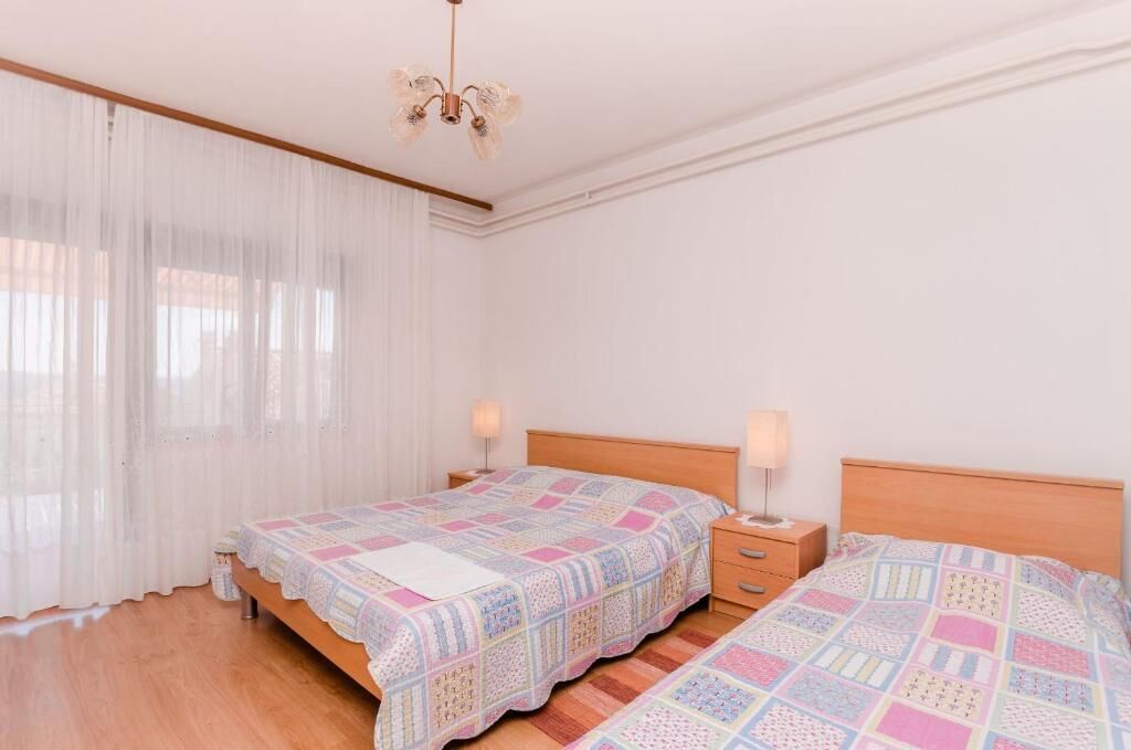Una camera da letto con due letti, una testiera in legno, un comodino con una lampada e tende trasparenti alla finestra.