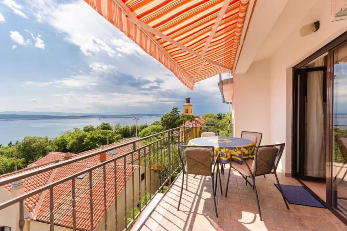 A balcony with a table and chairs, overlooking a body of water and a distant lighthouse.