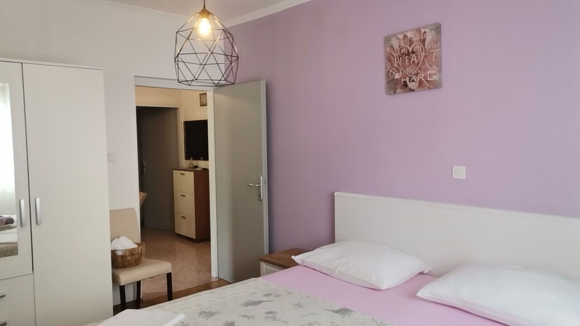 A bedroom with a bed, a bedside table, a hanging light fixture, a wardrobe, and a door leading to a bathroom.