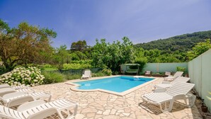 Une piscine dont le fond est recouvert de pierres, entourée de chaises longues blanches et d’une clôture verte.