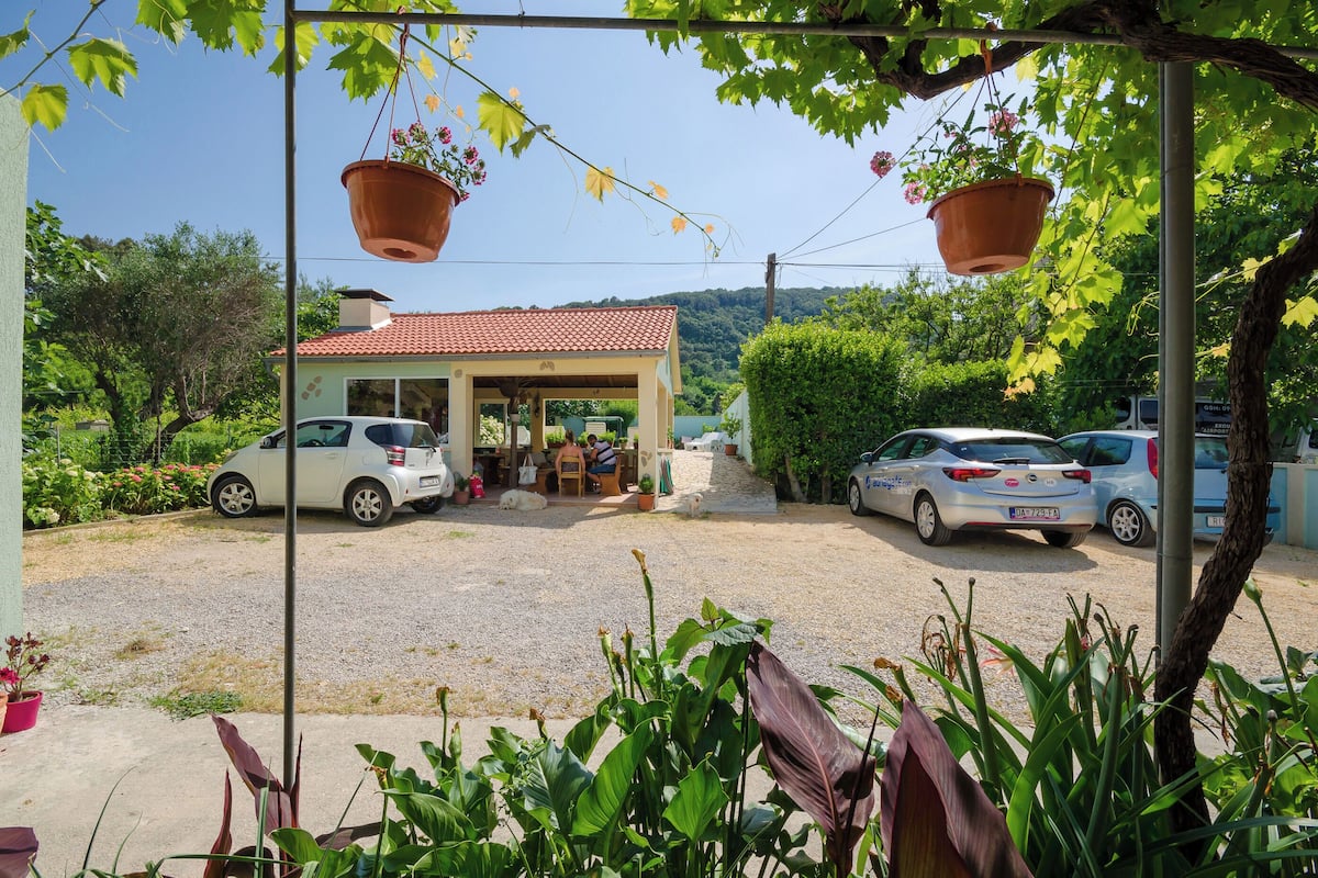 A house with a red-tiled roof, a covered porch, and a gravel driveway. There are two cars parked in front, and hanging flower pots are visible.