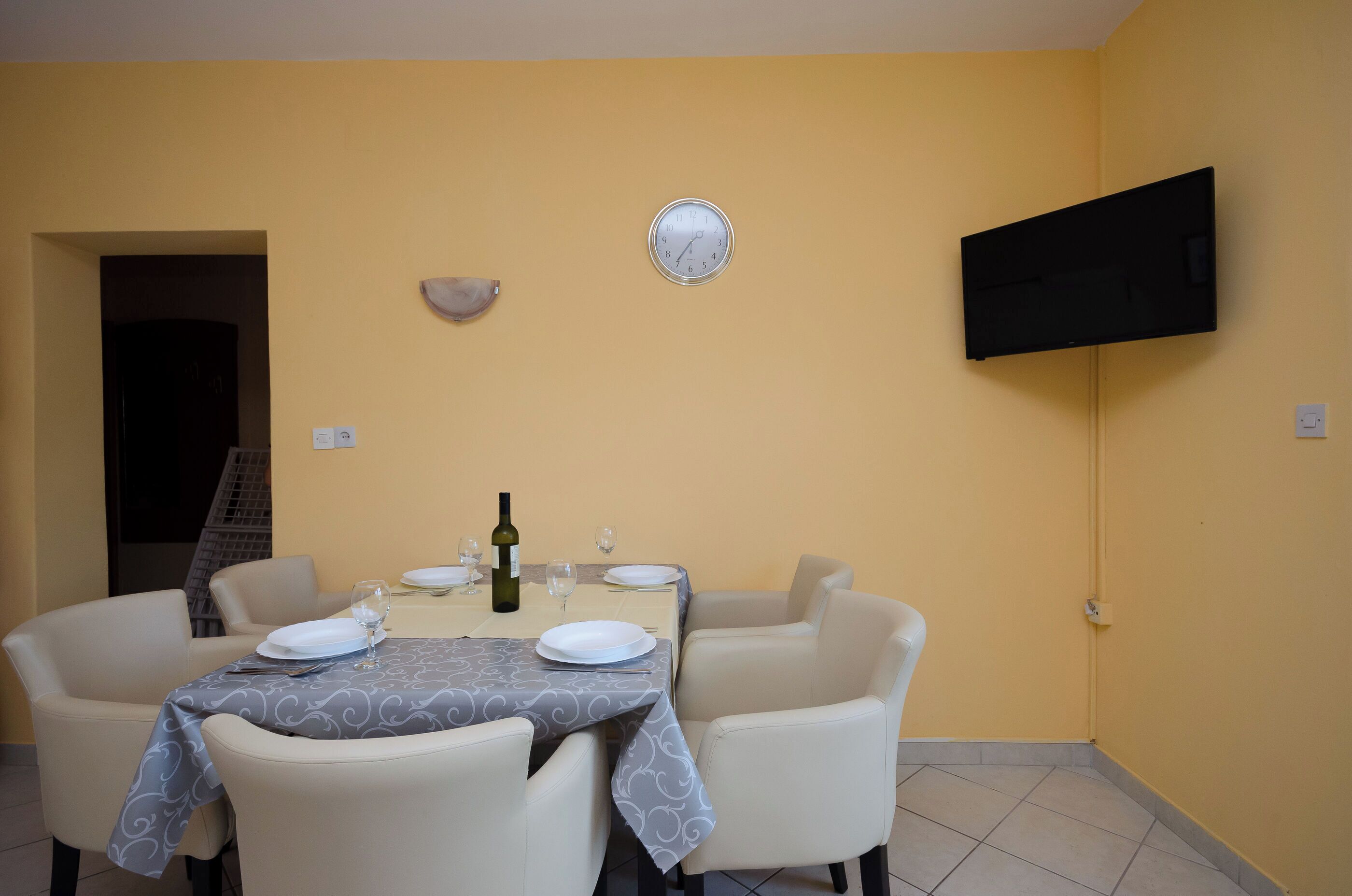 Comedor con una mesa dispuesta para cuatro, un reloj y un televisor de pantalla plana en la pared.