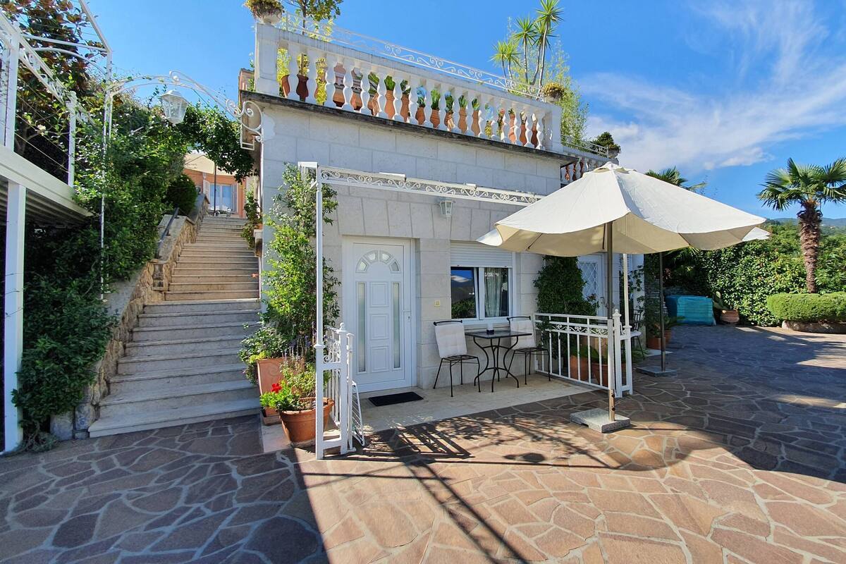 Eine Terrasse mit Steinboden, einer weißen Tür, einem kleinen Tisch mit Stühlen und einem großen Sonnenschirm.