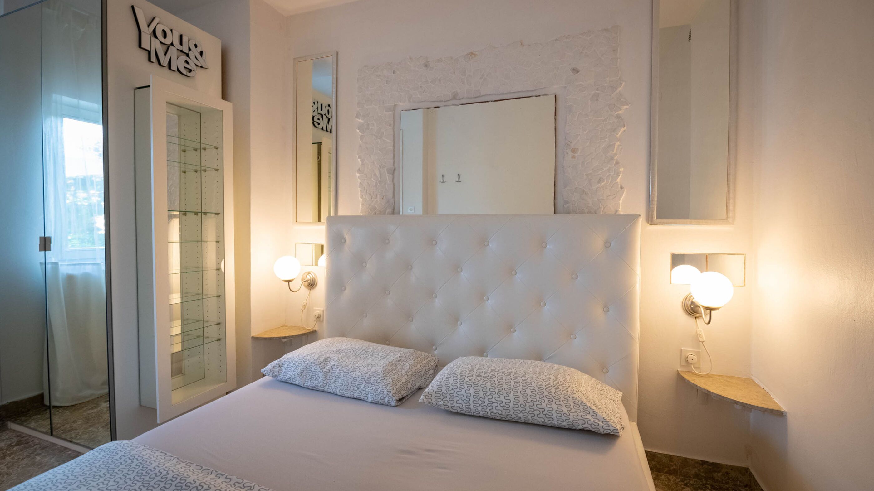 A bedroom with a large bed, two bedside tables with wall-mounted lamps, a mirror above the bed, and a glass door leading to a bathroom.