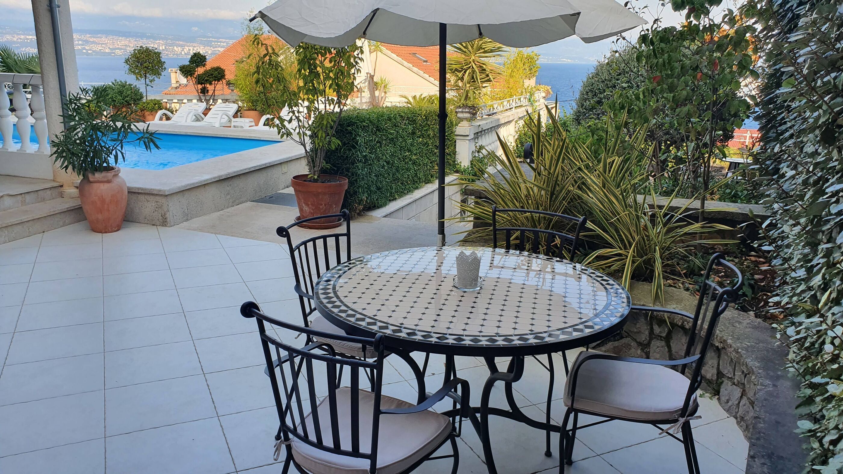 A round table with chairs and a white umbrella, surrounded by potted plants and a view of a swimming pool and buildings.