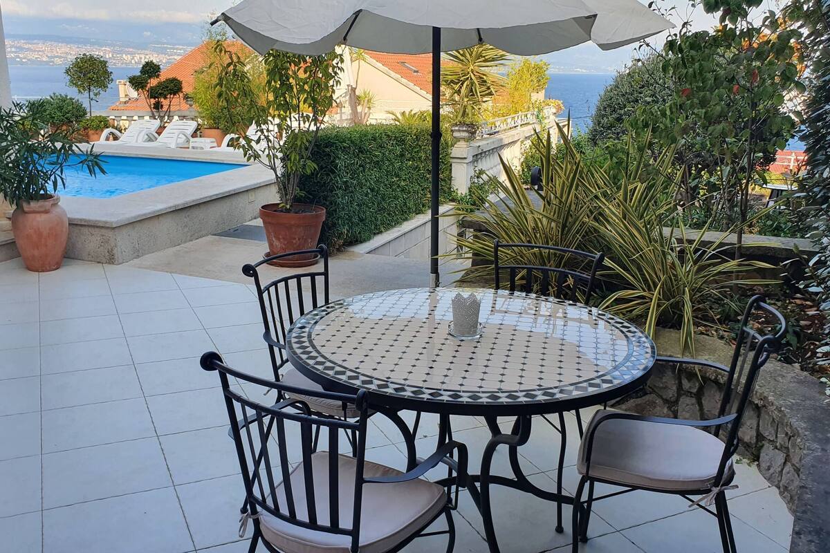 Ein runder Tisch mit Stühlen und einem weißen Sonnenschirm, umgeben von Topfpflanzen mit Blick auf einen Swimmingpool und Gebäude.