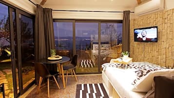 Quarto moderno com cama grande, TV, área de refeições com mesa e cadeiras, e vista para o exterior.