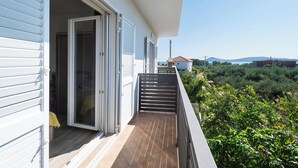 A balcony with a wooden floor, a glass door, and a view of a garden and distant buildings. - Apartment Summerteracea (Vodice)