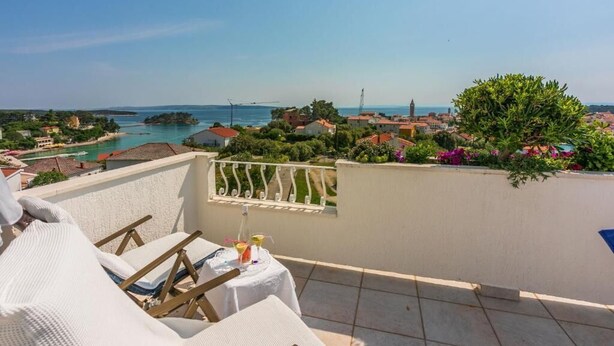 Un balcon avec une table dressée pour deux personnes, donnant sur une ville côtière avec ses bâtiments et un ciel dégagé.