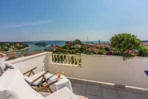 A balcony with a table set for two, overlooking a coastal town with buildings and a clear sky. - Apartments Vila Antula (Banjol)