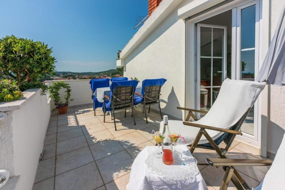 Una terraza con una mesa preparada para dos, una botella de bebida y una silla.
