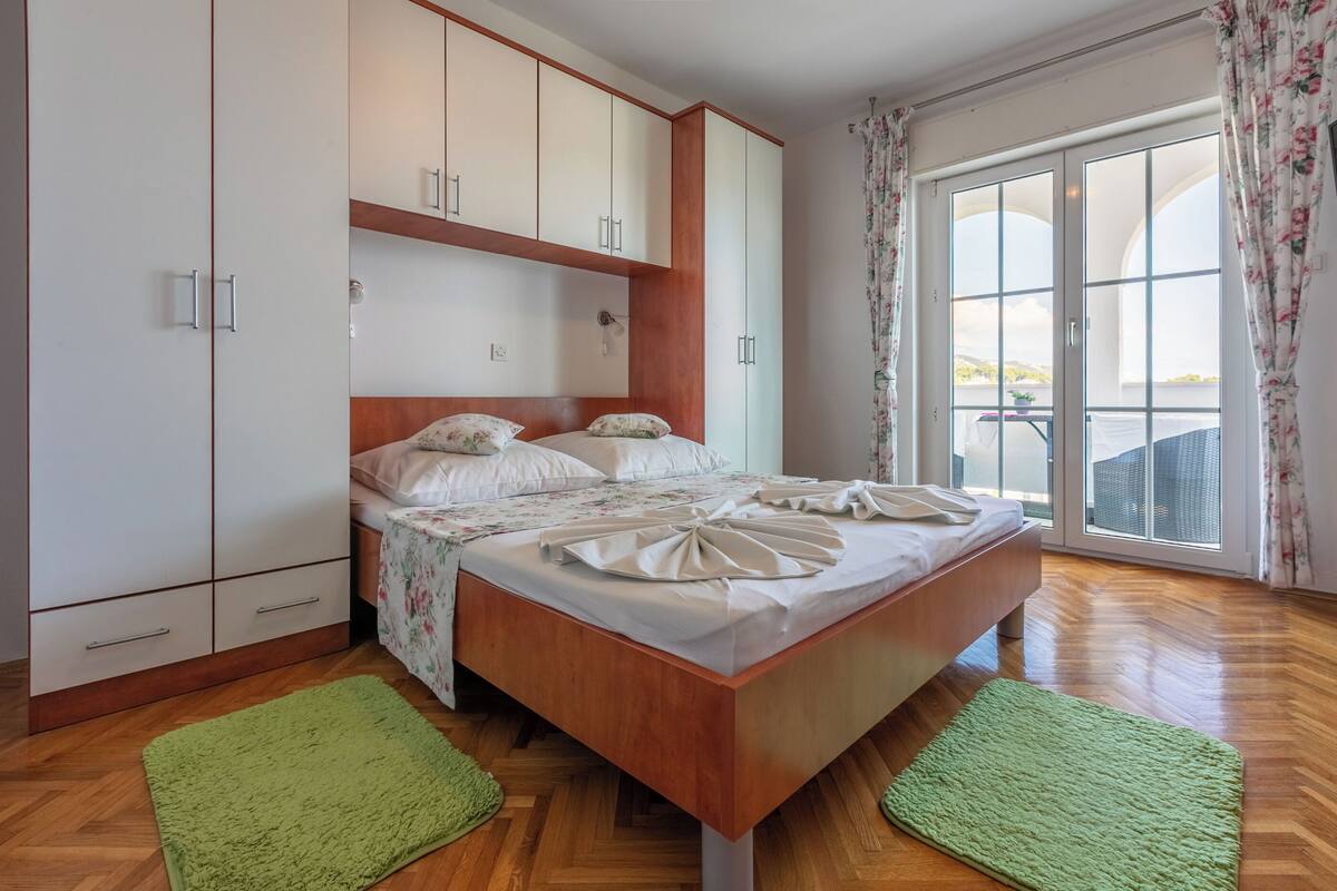 Un dormitorio con una cama de madera, ropa de cama blanca con motivos florales, una alfombra verde y un ventanal con cortinas.