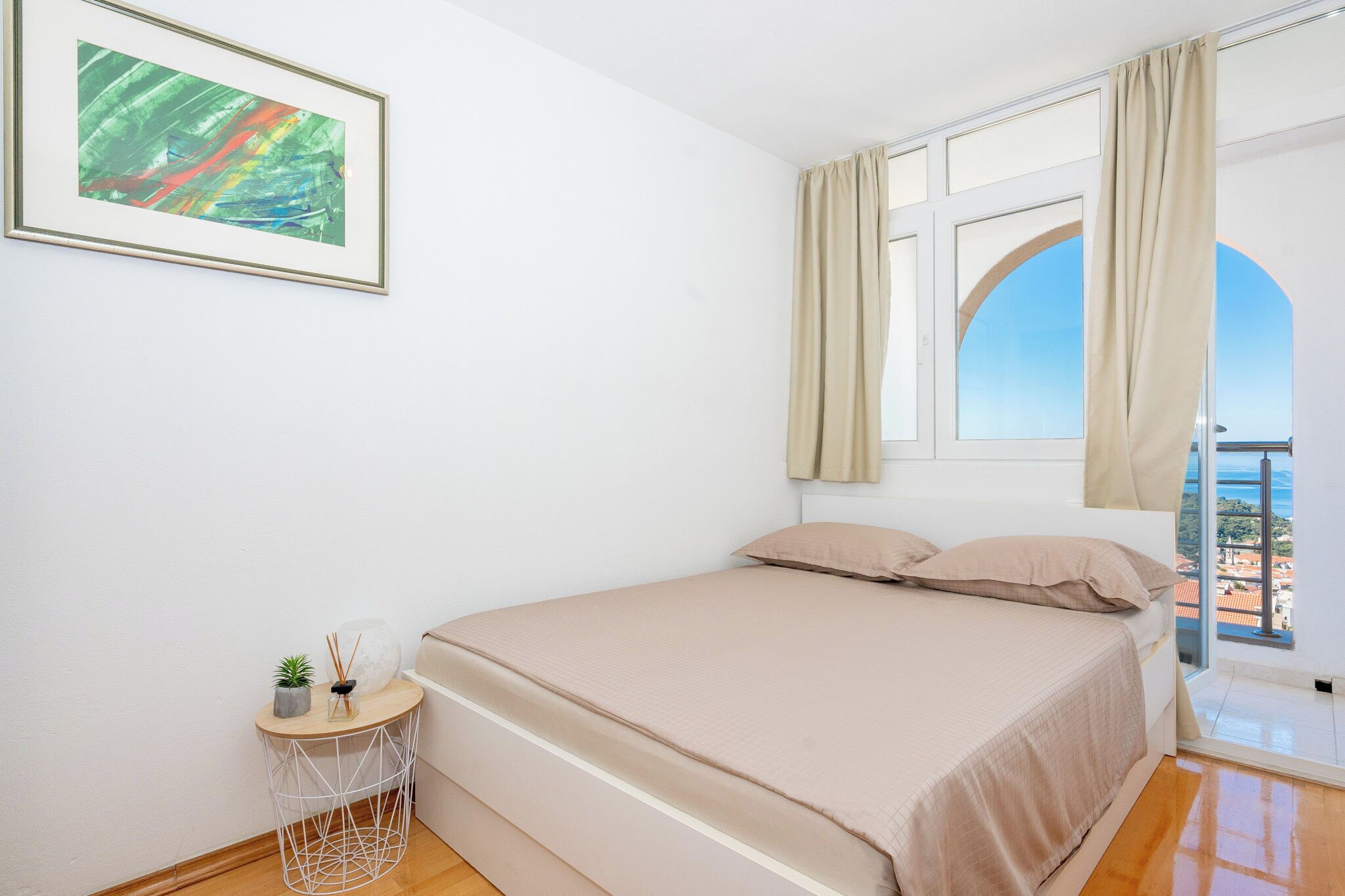 Ein Schlafzimmer mit einem Bett, einem kleinen Nachttisch, einem gerahmten Bild und einem Fenster mit Vorhängen.