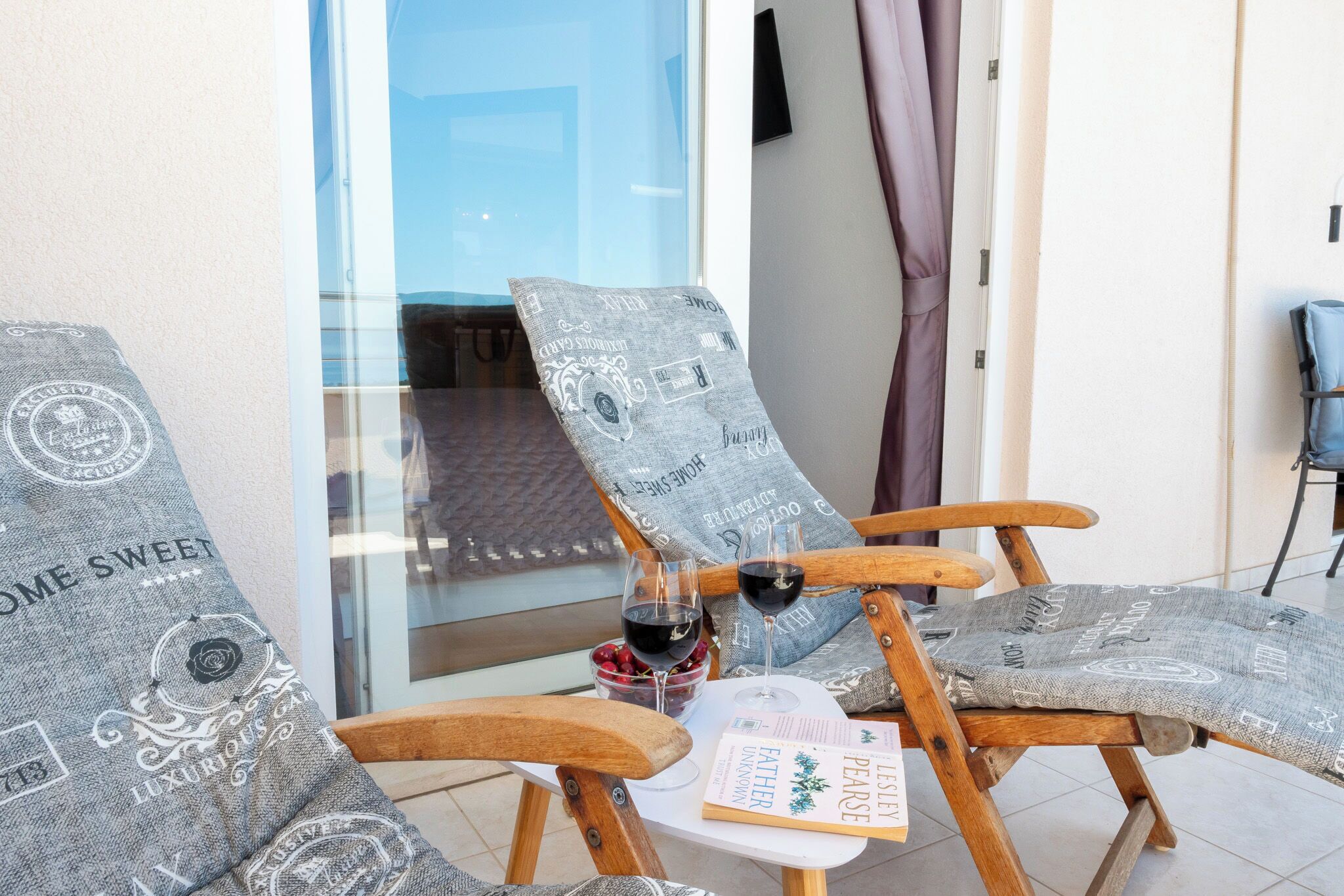 Two wooden chairs with grey patterned cushions, a glass table with a book and two wine glasses, and a glass bowl with fruit.