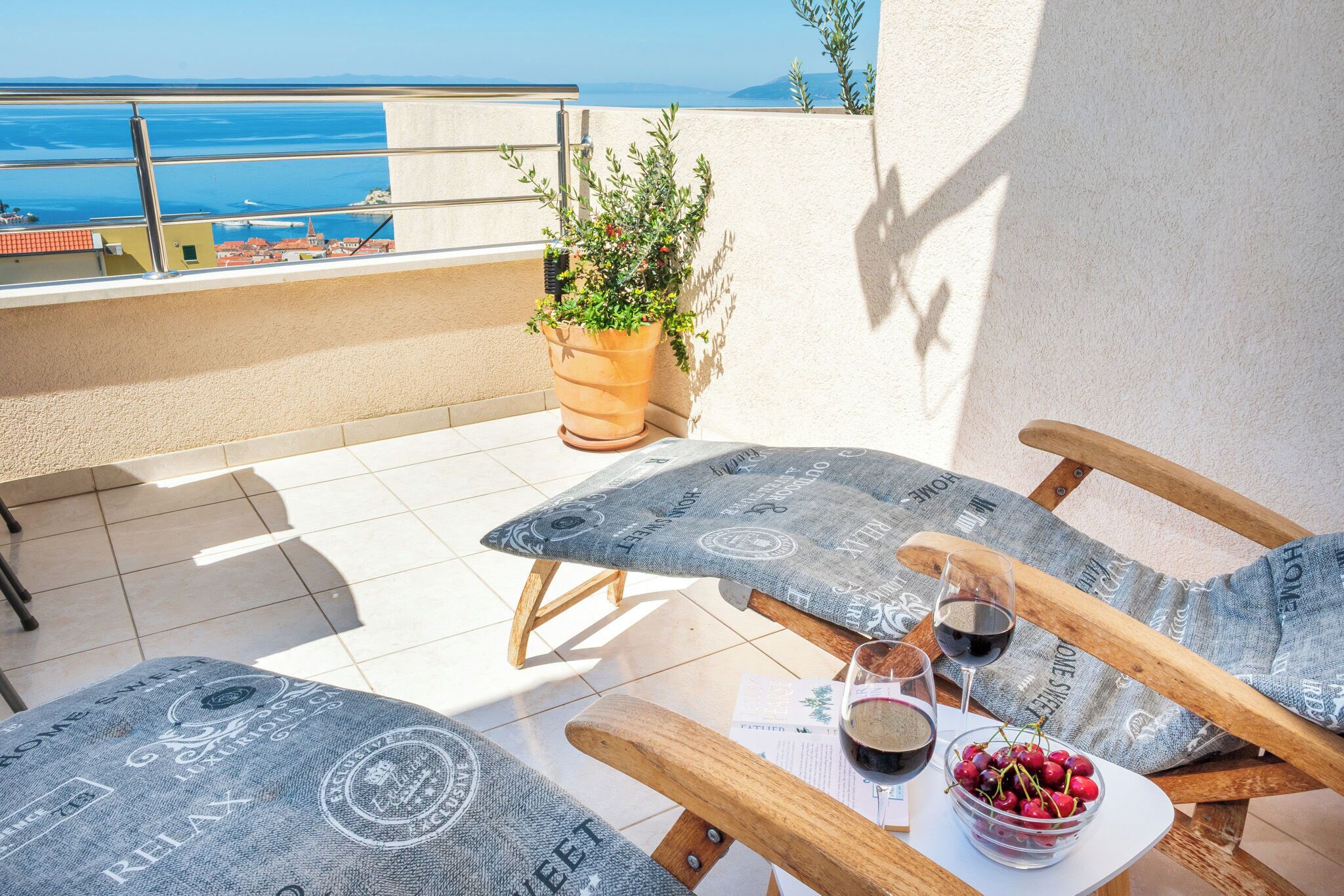 A terrace with two wooden lounge chairs, a table with a bowl of cherries and two glasses of red wine, and a potted plant.