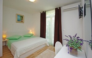 A bedroom with a bed, a nightstand, a lamp, a window with curtains, a wall-mounted air conditioner, and a vase with flowers. - Apartments Pavlinovic (Makarska)