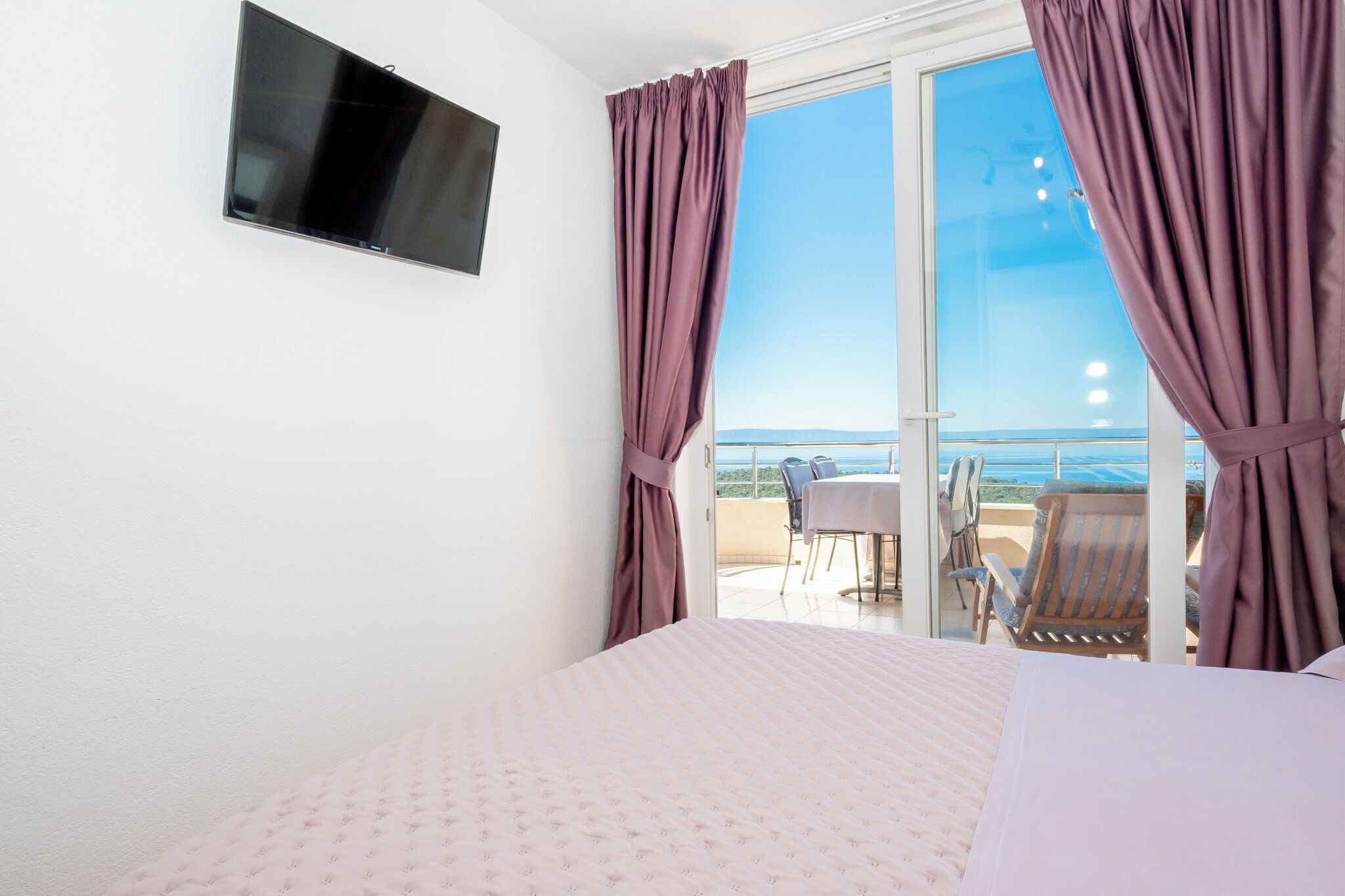 Een slaapkamer met een bed, een aan de muur bevestigde televisie en een balkon met een tafel en stoelen.