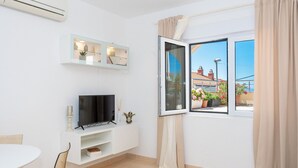 A modern living room with a TV, white walls, a window with curtains, and a balcony with potted plants. - Apartments Pavlinovic (Makarska)