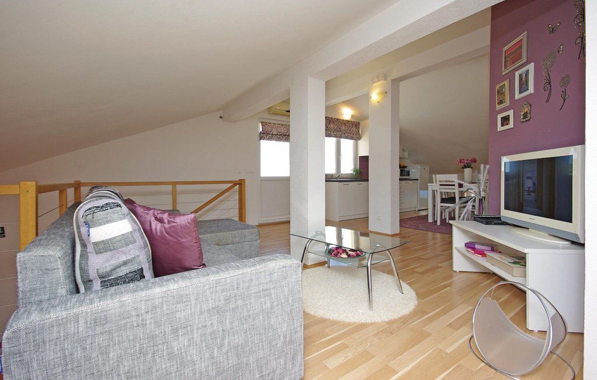 A modern apartment with a loft-style ceiling, a grey sofa with a purple pillow, a glass coffee table, a white TV stand, and a dining area visible in the background.
