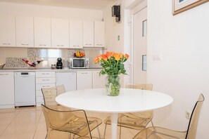 A modern kitchen with white cabinets, a microwave, and a dining area with a white table and clear chairs. - Apartments Pavlinovic (Makarska)