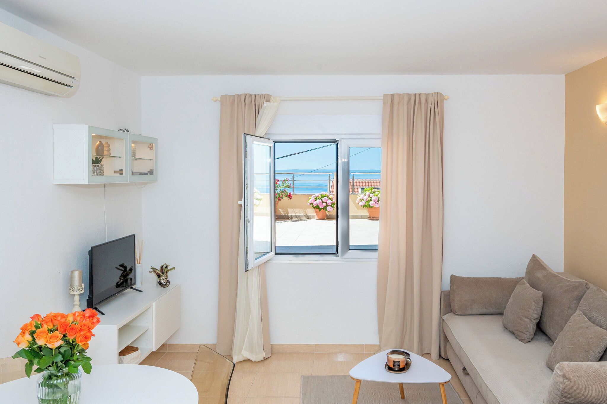 Une pièce avec une télévision, une petite table, un canapé, une fenêtre avec des rideaux et une vue sur la mer.
