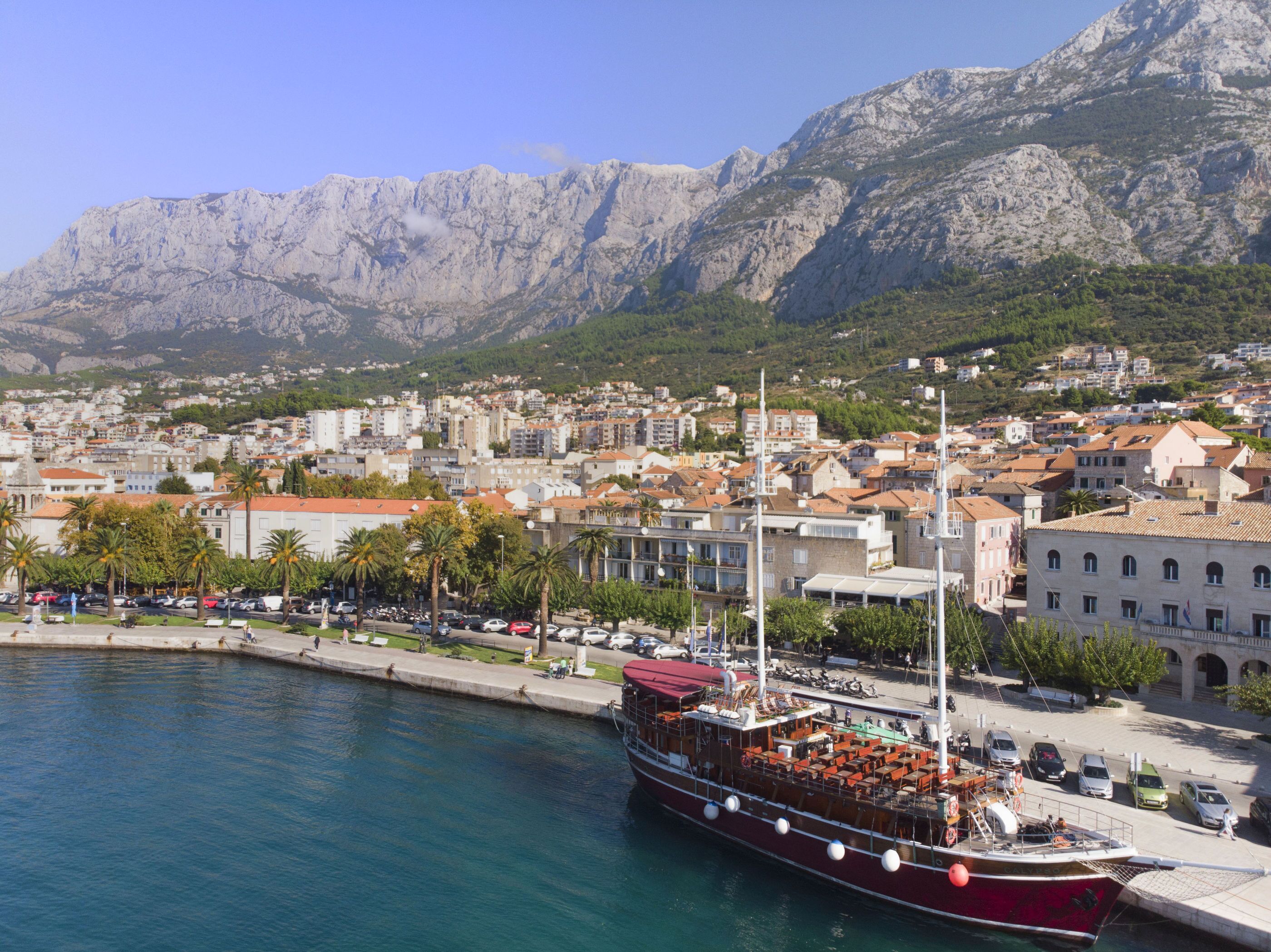 A coastal town with a prominent red boat docked at the pier, surrounded by buildings and mountains in the background. - Apartments Pavlinovic (Makarska)
