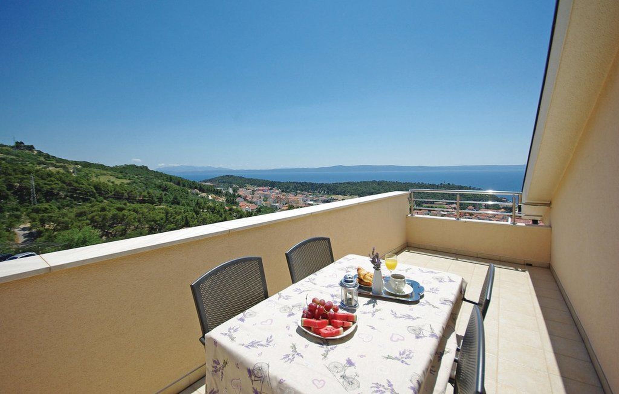 A balcony with a table set for two, overlooking a scenic view of a coastal town and hills.