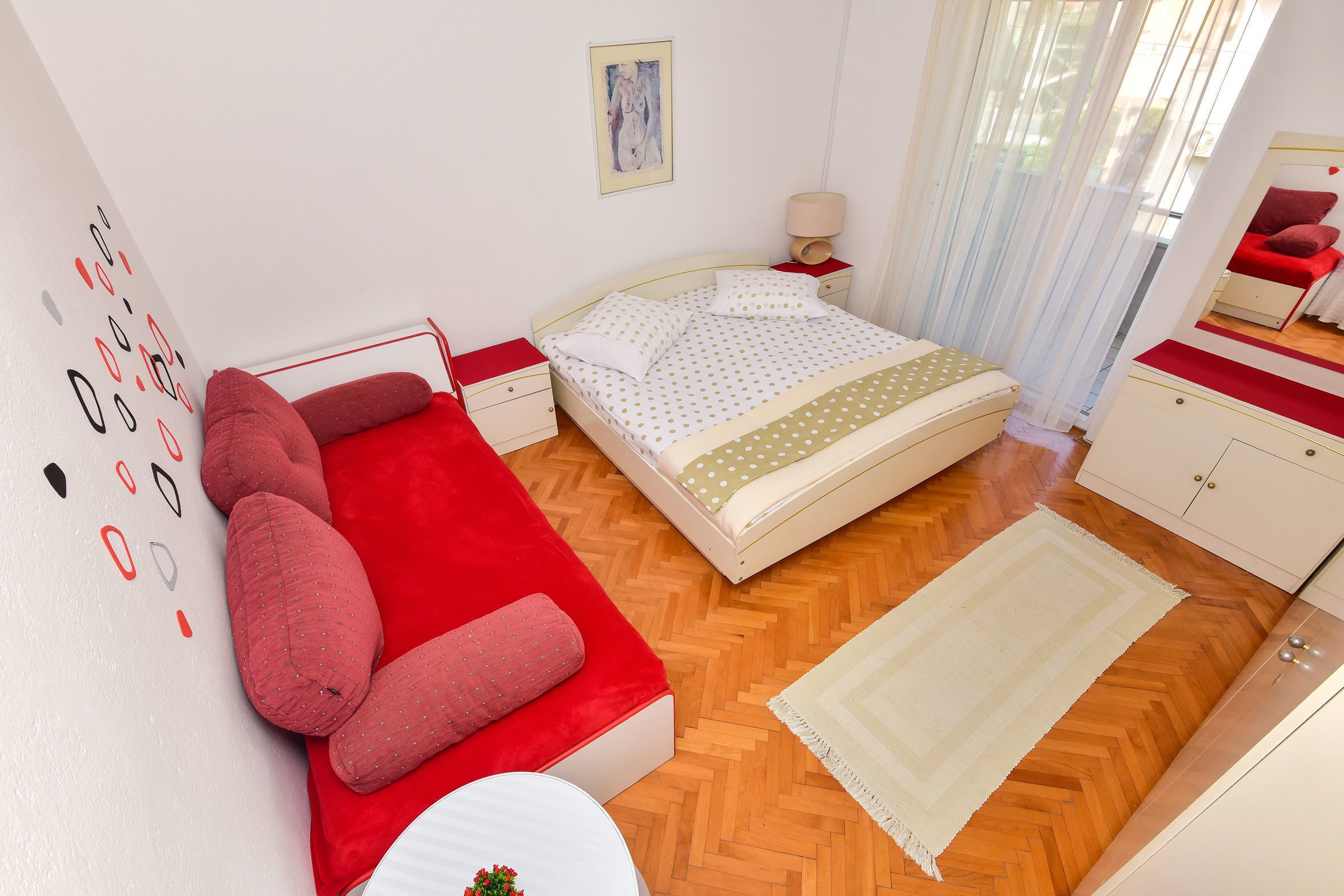 A bedroom with a bed, a red sofa, a nightstand, a lamp, and a painting on the wall.