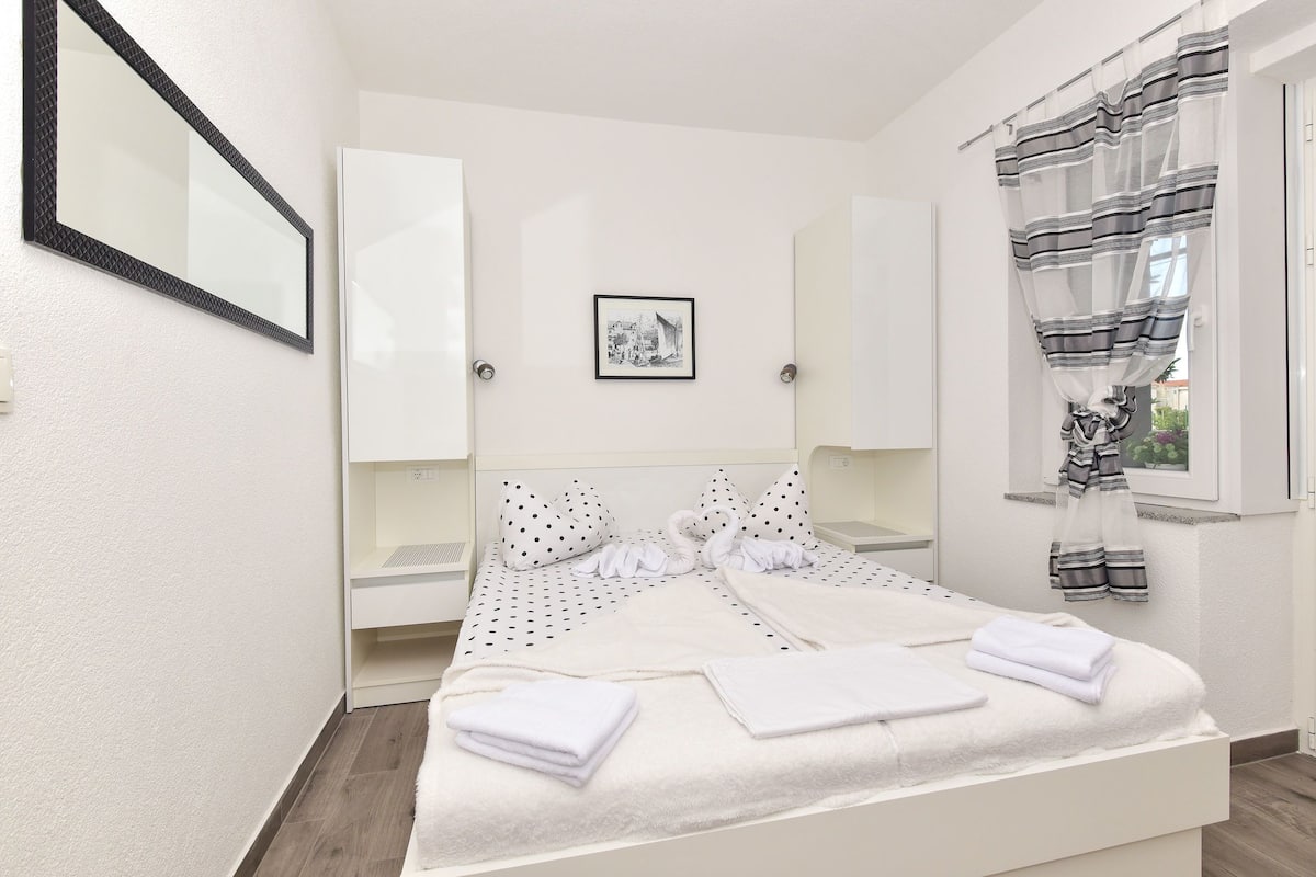 Un lit bien fait, avec du linge de lit et des serviettes blanches, un tableau encadré au mur et une fenêtre ornée de rideaux à rayures.