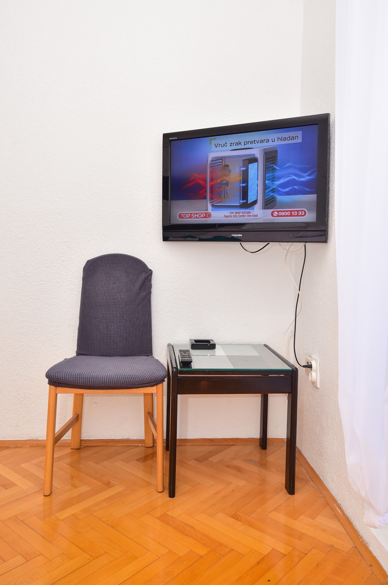 A room with a television mounted on the wall, a wooden chair, and a glass table.