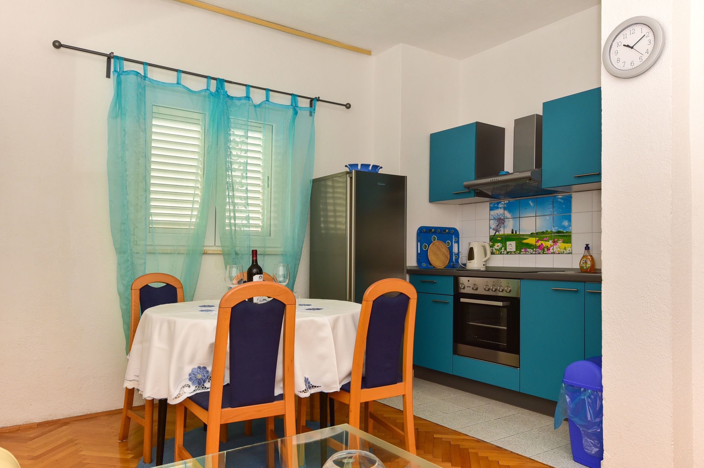 A compact kitchen with teal cabinets, a stainless steel refrigerator, and a small dining area with a glass table and wooden chairs.