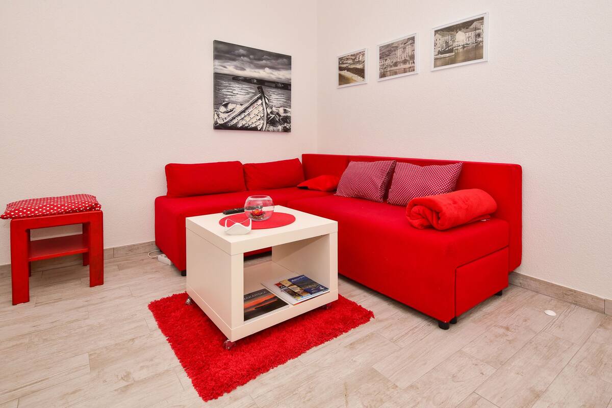 Un canapé rouge, une table d’appoint blanche, un tapis rouge et un tabouret rouge dans une pièce dont le sol est recouvert de parquet clair et les murs sont peints en blanc.