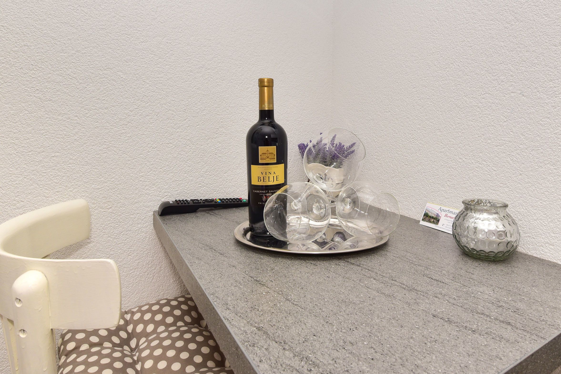 Una mesa con una botella de vino Vina Belje, un florero de cristal con flores moradas y un pequeño recipiente de cristal.