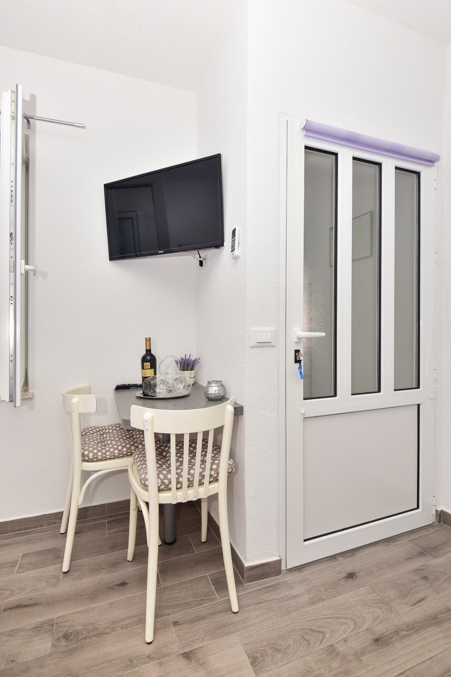 A room with a white door, a mounted TV, a dining table with chairs, and a bottle of wine.