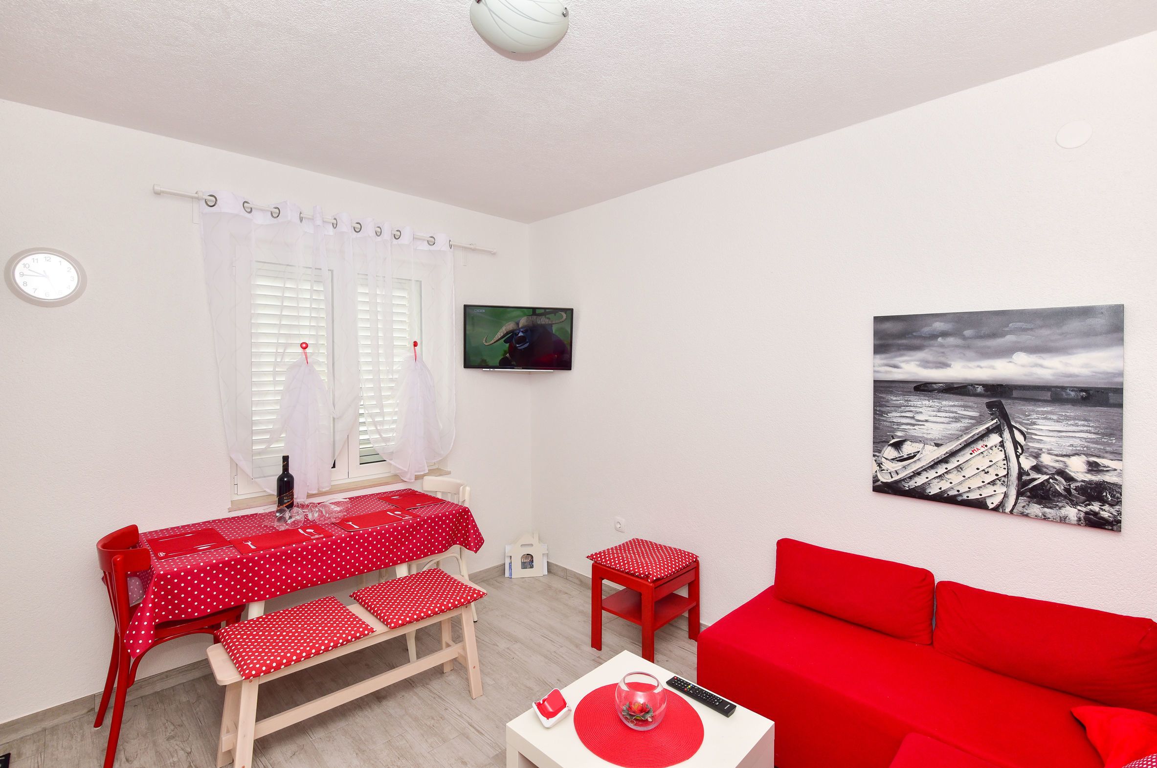 A room with a red table and chairs, a red sofa, a painting of a boat, and a window with curtains.