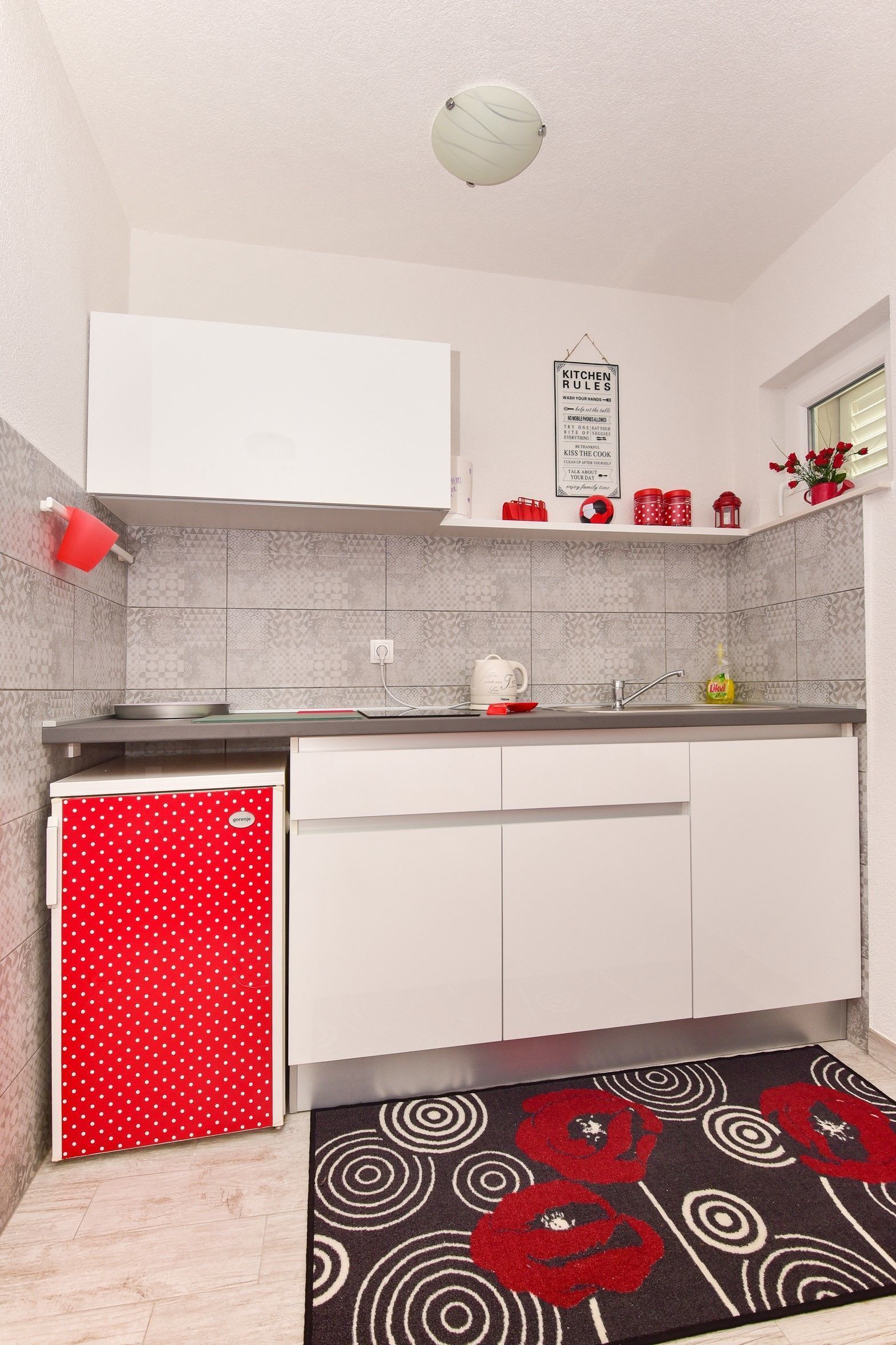Una cocina con armarios blancos, un refrigerador rojo con lunares, un tapete negro con flores rojas y un cartel que dice 'Kitchen Rules'.