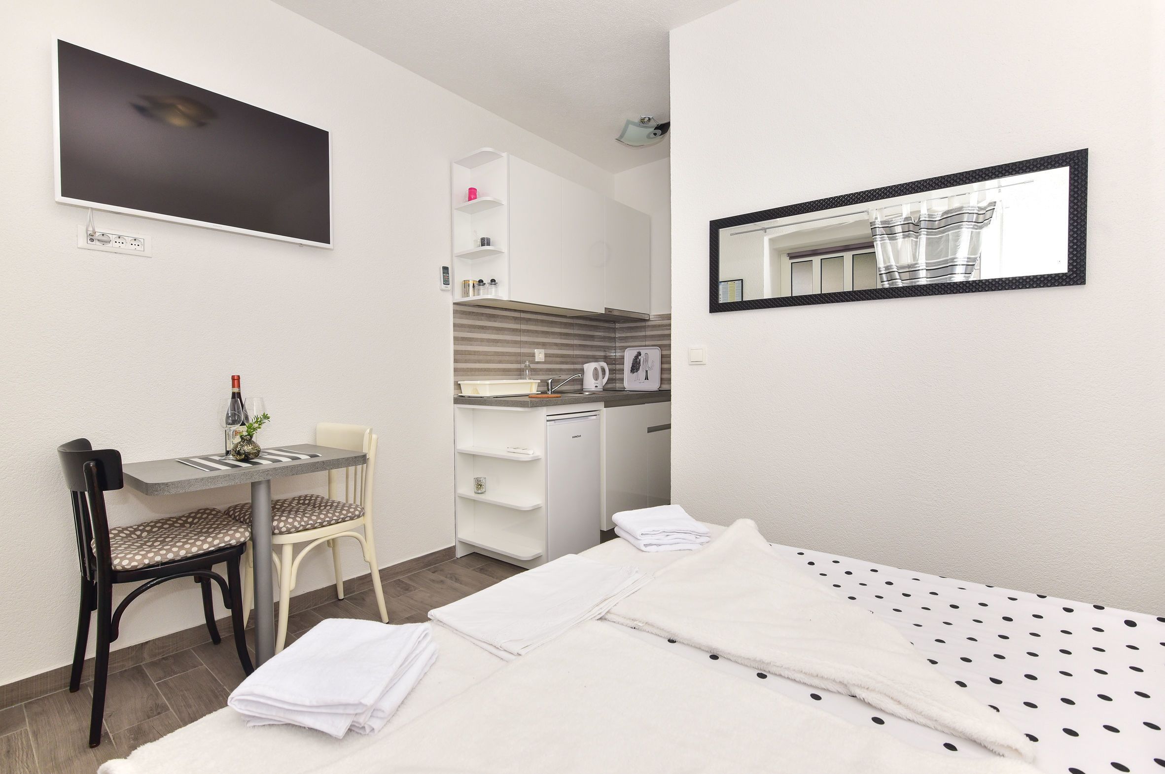 A compact hotel room with a bed, a desk with a chair, a small kitchenette, and a mirror.