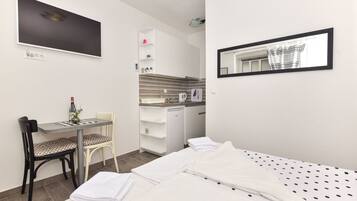 A compact hotel room with a bed, a desk with a chair, a small kitchenette, and a mirror.