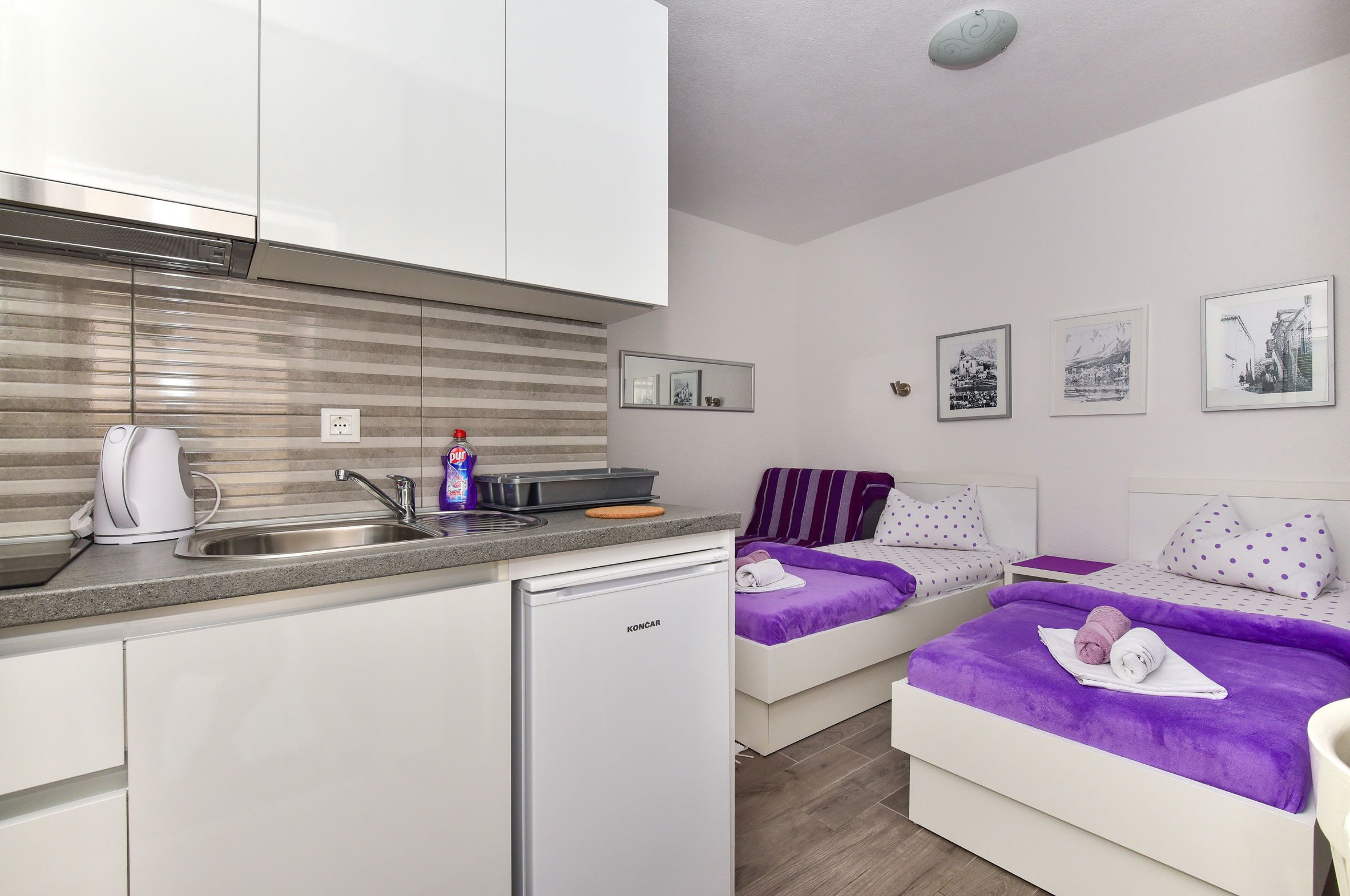 A compact hotel room with a kitchenette, two beds with purple bedding, and a small dining area.