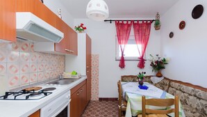 A compact kitchen with a gas stove, sink, and wooden cabinets. There is a dining area with a table and chairs, and a window with red curtains. - Apartment Anica (Kampor)
